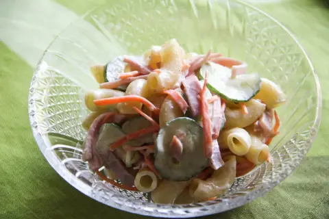 A close-up of a crystal bowl filled with creamy Japanese macaroni salad, featuring elbow macaroni, shredded carrots, sliced cucumbers, and ham.