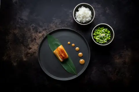 A perfectly cooked nobu miso black cod rests on a dark plate with a green leaf. Drizzled sauce and sides of rice and edamame complete the elegant dish.