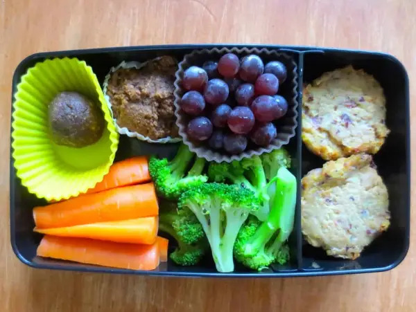 A bento box packed with bento nutrition: a date ball, muffin, grapes, carrots, broccoli, and two savory patties.